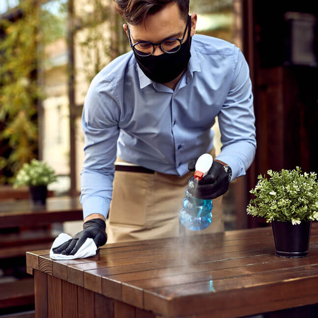 Restaurant employee wearing mask and cleaning off table with antibacterial spray
